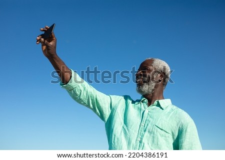 Front high angle view of a senior African American man standing on the beach with blue sky in the background, smiling and taking selfie