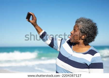 Side view close up of a senior African American woman standing on the beach with blue sky in the background, making a funny face and taking selfies with a smartphone 