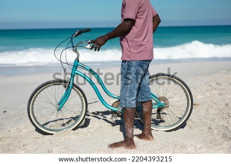 Low section of a senior African American man standing barefoot on the beach with blue sky and sea in the background, holding a blue bicycle  Royalty-Free Stock Photo #2204393215