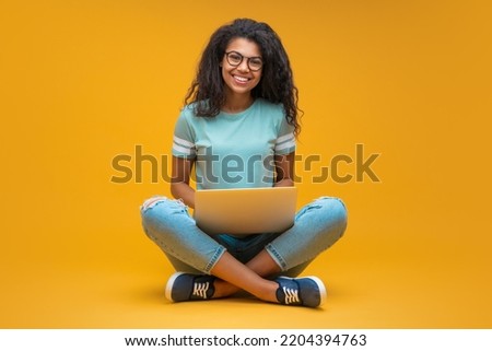 Studio portrait of smiling young beautiful african american girl in trendy spectacles, sitting with laptop and looking directly to the camera, isolated over bright orange yellow background.