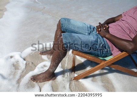 Low section side view of a senior African American man on a beach, sitting in a deckchair, resting with the sea lapping around his feet Royalty-Free Stock Photo #2204395995