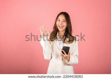 Happy Asian portrait beautiful cute young woman excited holding mobile phone and gesturing ok sign for agree, studio shot isolated on pink background, Thai female making finger symbol on smartphone