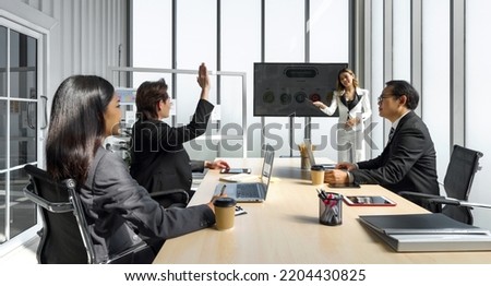 Young asian businessman in suit raise his hand to ask a question to presenter standing in front of a  large digital monitor. Business executives team meeting in modern office.