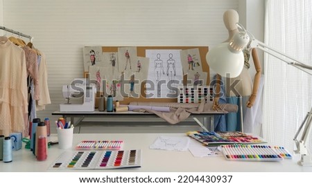 Fashion designer room. Sewing Machine and various sewing related items on the table. Mannequins standing, colorful fabrics and a clothes rack with various kind of dress place beside the table.
