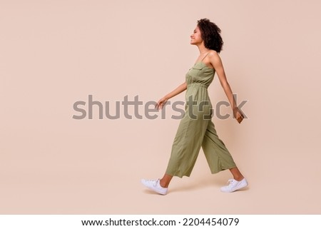Side profile photo of cheerful nice woman with wavy hairdo dressed khaki overall white sneakers walking isolated on beige color background Royalty-Free Stock Photo #2204454079