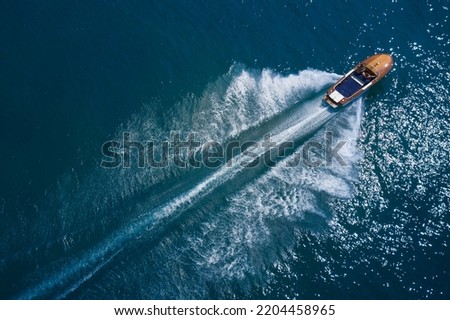 Luxurious wooden boat fast movement on dark water. Luxurious wooden motor boat rushes through the waves of the blue Sea. Classic Italian wooden boat fast moving aerial view. Royalty-Free Stock Photo #2204458965
