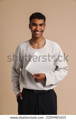 Portrait of young handsome smiling african man with hand in pocket holding laptop and looking at camera , while standing over isolated beige background Royalty-Free Stock Photo #2204481655