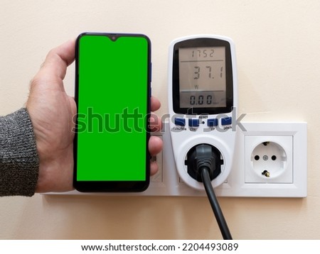 A man holds a smartphone with a green screen next to an electricity consumption meter near a power outlet. Analysis of electricity consumption using the phone.