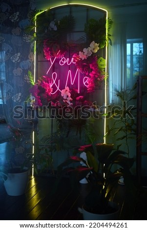 Beautiful bright neon letters with tropical flowers. Words you and me. Orchids and palm leaves. Abstraction. Romance and love. Bright colors. Reflection in the mirror. Dark interior