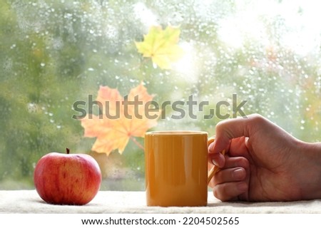 yellow mug in a hand and a red apple on the background of a window after the rain. autumn breakfast with hot drink