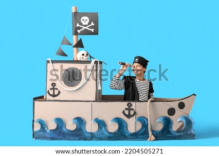 Little boy dressed as pirate with spyglass in cardboard ship on blue background Royalty-Free Stock Photo #2204505271