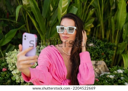 Young beautiful brunette in a pink shirt, neck jewelry, necklace, trendy sunglasses sits on a background of tropical leaves, takes a selfie on the phone