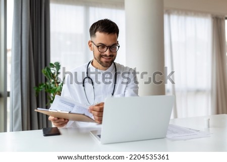 Friendly young doctor uses laptop to remotely communicate with patient, man in clinic consults online, looks at monitor screen and webcam