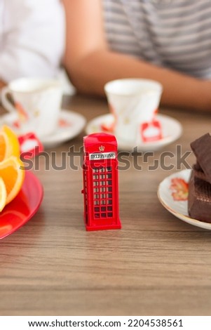 Traditional afternoon tea of british ceremony with such symbol of britishness as toy telephone box, defocused faceless women on background, front focus, vertical image