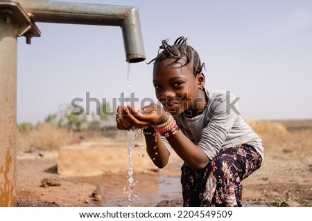 Smiling african girl tasting fresh water flowing from a tap in the middle of an arid rural landscape, withelandscape; access to clean water and sanitation concept Royalty-Free Stock Photo #2204549509