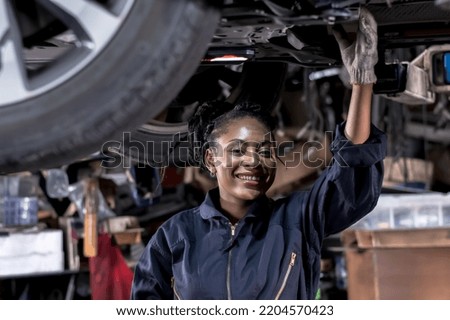 Smiling African American black mechanic woman in overall working underneath lifted vehicle in car repair shop,happy black car female mechanic fixing vehicle,car maintenance and auto repair concept Royalty-Free Stock Photo #2204570423