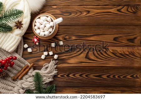 Christmas Eve concept. Top view photo of hat knitted scarf pine branches mug of cocoa with marshmallow on rattan placemat viburnum berries and cinnamon sticks on wooden table background with copyspace