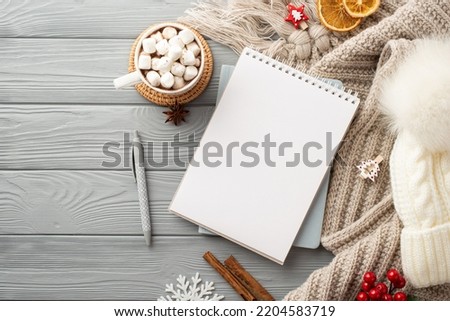 Top view photo of notepad pen bobble hat plaid cup of hot drinking with marshmallow serving mat snowflake cinnamon sticks viburnum and dried orange slices on grey wooden desk background with copyspace