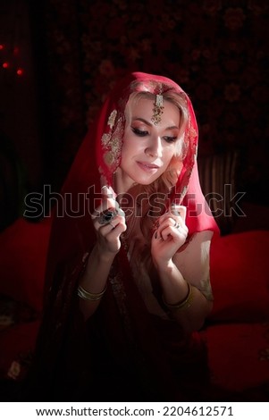 A beautiful European girl looking like an Arab woman in a red room in a harem. Photo shoot of an oriental style odalisque. A model poses in a sari as an indian woman in india