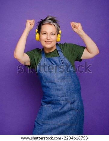 Beautiful mature woman in denim apron with yellow headphones listens favorite music and dancing isolated over violet background. Dancing mum while cooking. Grey haired cheerful woman love dancing