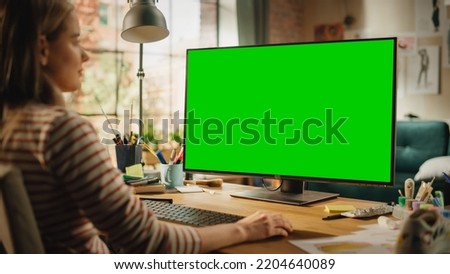 Over the Shoulder View of Creative Young Woman Sitting at Her Desk, Using Desktop Computer with Mock-up Green Screen. Female Caucasian Specialist Working on Computer with Chroma Key Display at Home.