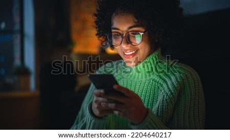 Cinematic Portrait of Black Female with Curly Hair Sitting on the Floor at Home and Using Smartphone. Diverse Woman Receives Good News and Chatting with Friends Via Social Media. Royalty-Free Stock Photo #2204640217