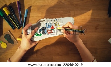 Top View Shot: Designer Sitting Behind the Wooden Desk at Home and Applying Colourful Pictures on Shoes. Hand Made Stylish Footwear and Creative Process Concept. Close Up.