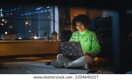 Positive Latina Female Using Laptop with Stickers at Home. Woman Resting on the Floor and Messaging on Computer with Smile on Face. Evening City View Behind the Big Window. Royalty-Free Stock Photo #2204640227