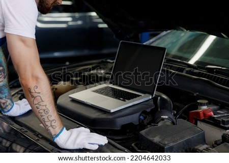Close up cropped repairman technician car mechanic man arms hand typing use laptop pc computer make doing diagnostics check fix problem with raised hood bonnet working in vehicle repair shop workshop.