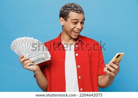 Young surprised excited man of African American ethnicity he wear red shirt hold in hand fan of cash money in dollar banknotes use mobile cell phone isolated on plain pastel light blue cyan background