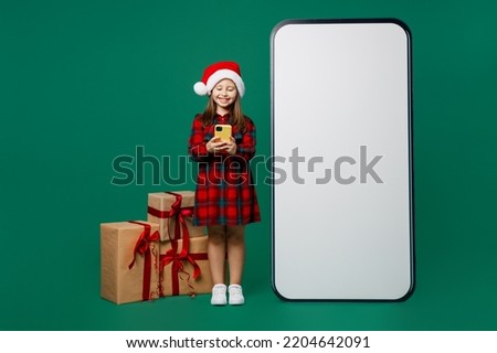 Full body little child kid girl 7 years old wear red dress Christmas hat posing big blank screen mobile phone with area use smartphone isolated on plain green background Happy New Year 2023 concept
