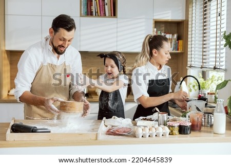 Cooking preparing process. Father sieving flour on wooden surface young daughter sprinkling helping dad. Young mom preparing pizza ingredients eggs salami butter mushrooms on table.