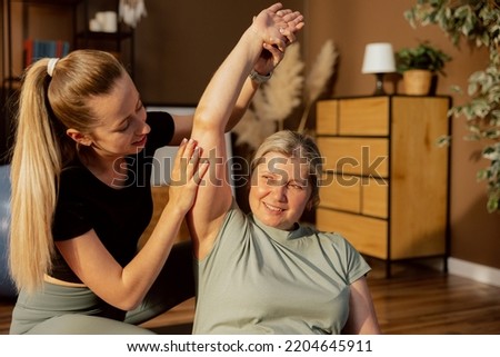 Delighted young nurse and senior woman indoors doing exercises. Young lady helping elderly woman raising hands stretching breathing relaxing.