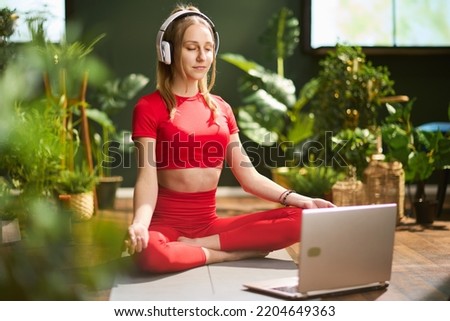 young female in red fitness clothes with laptop and headphones having webcam meeting and meditating in the modern green living room. Royalty-Free Stock Photo #2204649363
