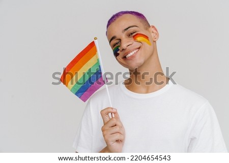 Portrait of young handsome stylish smiling happy boy with LGBT flag painted on his cheek, holding LGBT flag in his hand and looking at camera , while standing over isolated white background Royalty-Free Stock Photo #2204654543