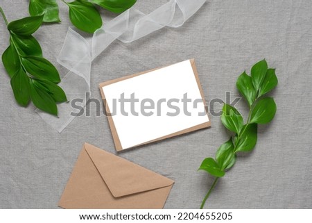 Blank invitation wedding card mockup, brown envelopes, ribbon and green leaves. Grey linen textile background. Top view, flat lay.