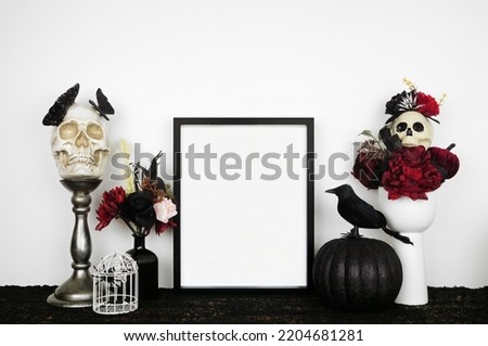 Halloween gothic romance mock up. Black frame on a black shelf with red and black flowers, skulls and pumpkin. Portrait frame against a white wall. Copy space. Royalty-Free Stock Photo #2204681281