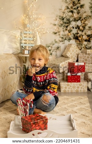 Cute blond toddler preschool boy, eating cookies and opening present on Christmas on cozy home, lights and decoration