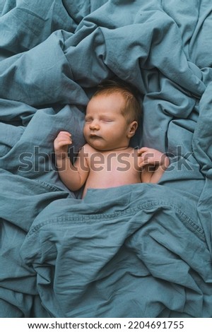 White newborn baby boy sleeping in blue bedsheets with arms up and curled fingers looking cozy and calm. First baby photoshoot. Vertical shot . High quality photo