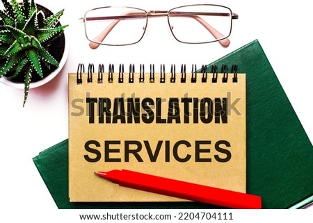 On a light background, gold-framed glasses, a flower in a pot, a green notebook, a red pen and a brown notebook with the text TRANSLATION SERVICES. Business concept