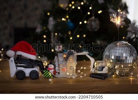 models of toy excavator and loader, souvenir snowman, house with candle, sparkler standing in mirror ball against of Christmas tree. Concept of Christmas business greetings for construction companies