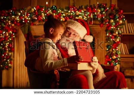 Side view portrait of cute African American girl opening Christmas present while sitting on Santas lap
