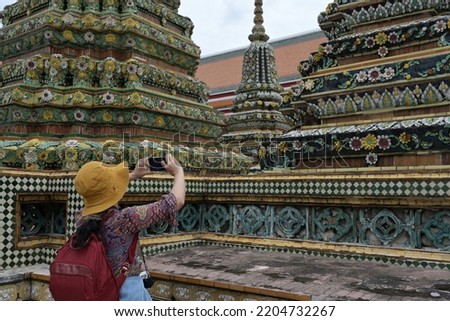 A female traveler wearing a yellow hat with a red backpack and jeans is using mobile phone. Take photos of the magnificent pagodas in famous temples: Wat Pho or the Reclining Buddha Temple in Bangkok