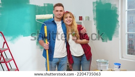 Young Caucasian joyful positive married couple standing in new own apartment holding paint brushes during home repair, hugging, looking at camera and smiling, home renovation, house repair concept