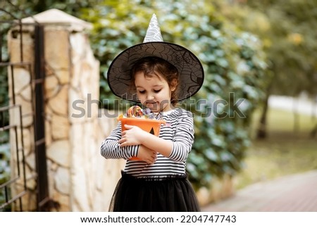 Little cute girl in witch costume holding jack-o-lantern pumpkin bucket with candies and sweets. Kid trick or treating in Halloween holiday