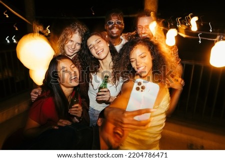 A cheerful group of friends are taking selfies at the rooftop party. They are smiling, making faces, and posing. Memories during summertime at the open-air club. Royalty-Free Stock Photo #2204786471