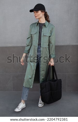 A beautiful girl is wearing a grey t-shirt, cloth cap, coat, black bag and jeans posing against the background of a wall. fashion outfit for the city, minimalism urban style clothing
