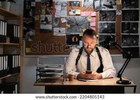 male detective is sitting in the office flipping through the news feed. A detective board with photos, a map and clues connected by a thread on the wall. copy space.