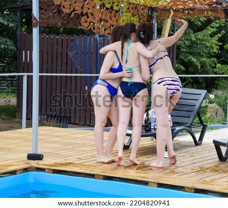 Three girls in swimsuits take a selfie standing by the pool. Beautiful female buttocks, stand by the sun lounger and take pictures.