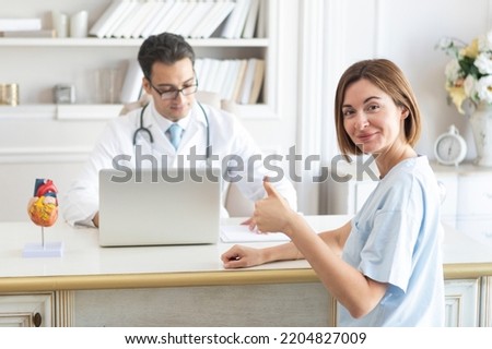 Portrait of young cheerful female patient showing thumbs up and doctor cardiologist working in office of modern clinic	 Royalty-Free Stock Photo #2204827009